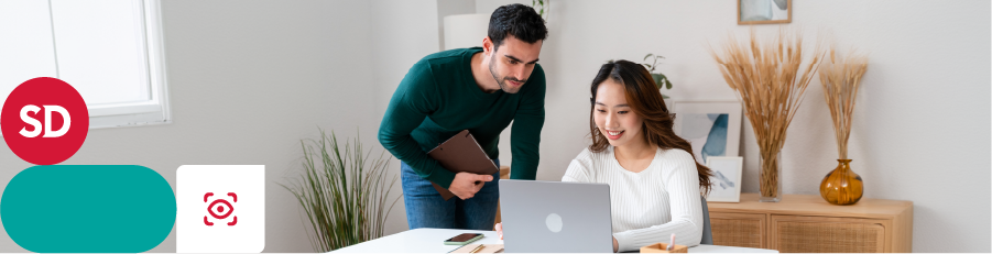 Two individuals collaborating, one seated with a laptop and another standing with a tablet in a modern office.

