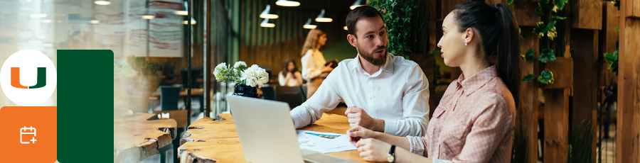 "Two people discussing work at a table in a modern café with a laptop and documents."